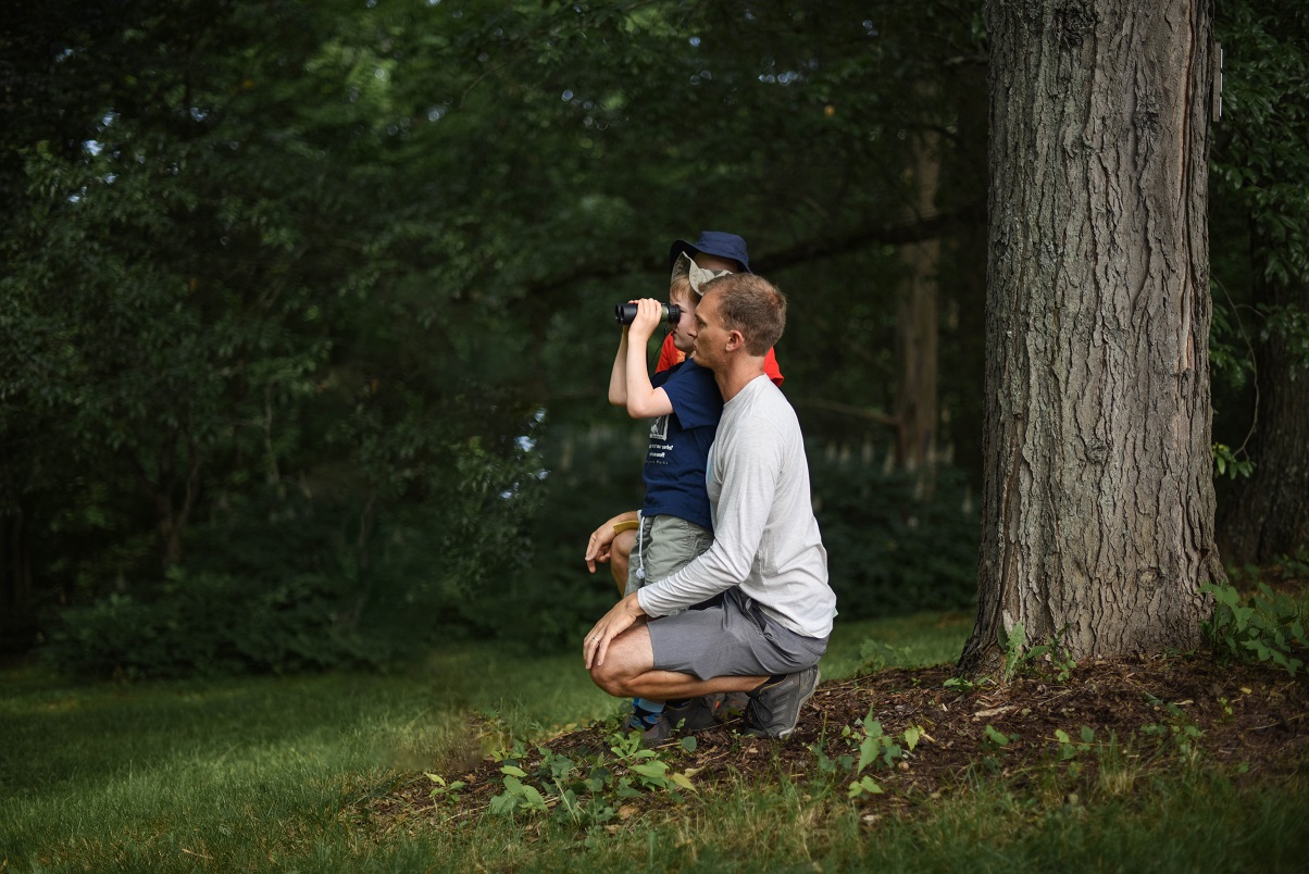 IMage of father looking over the shoulder while his son is looking through binnoculars.