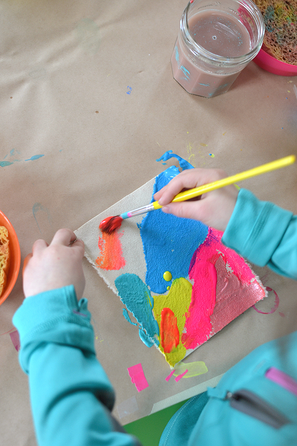 A child's hands painting a small canvas using orange, blue, and pink paints.