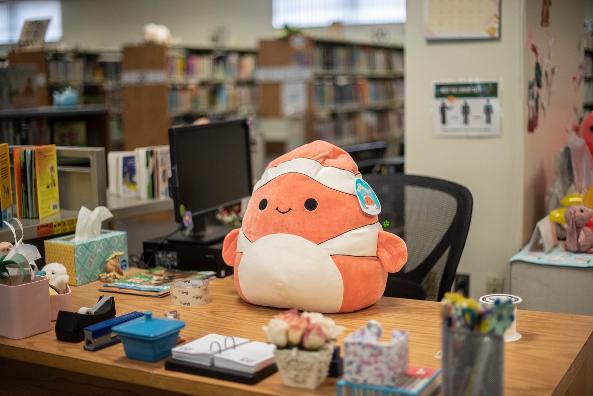 A clown fish Squishmallow is perched ontop of a desk.