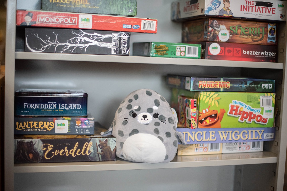 A spotted seal squishmallow on a shelf with boardgames.