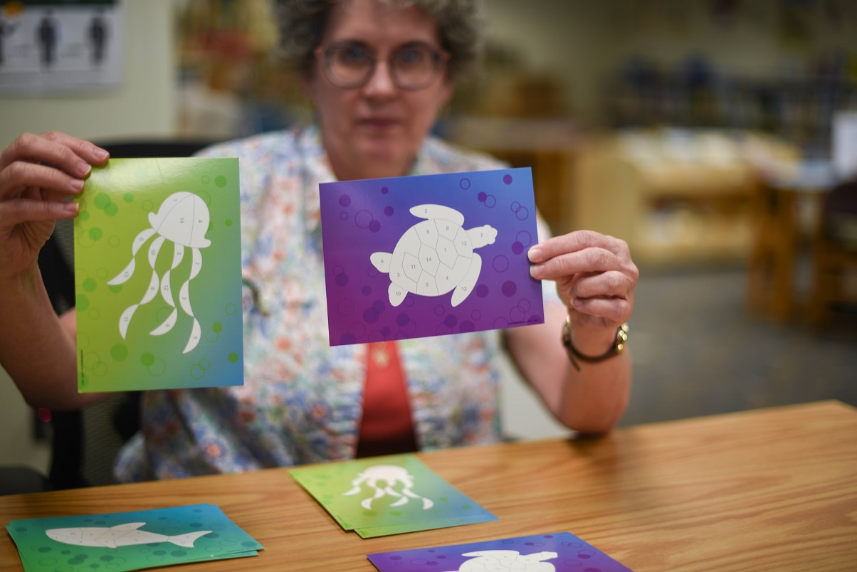 Ruth, children's librarian, is holding color by numbers paper images of sea creatures like turtles and jellyfish